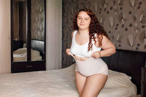Plus-size woman adjusting her comfortable nude shapewear in a bedroom.